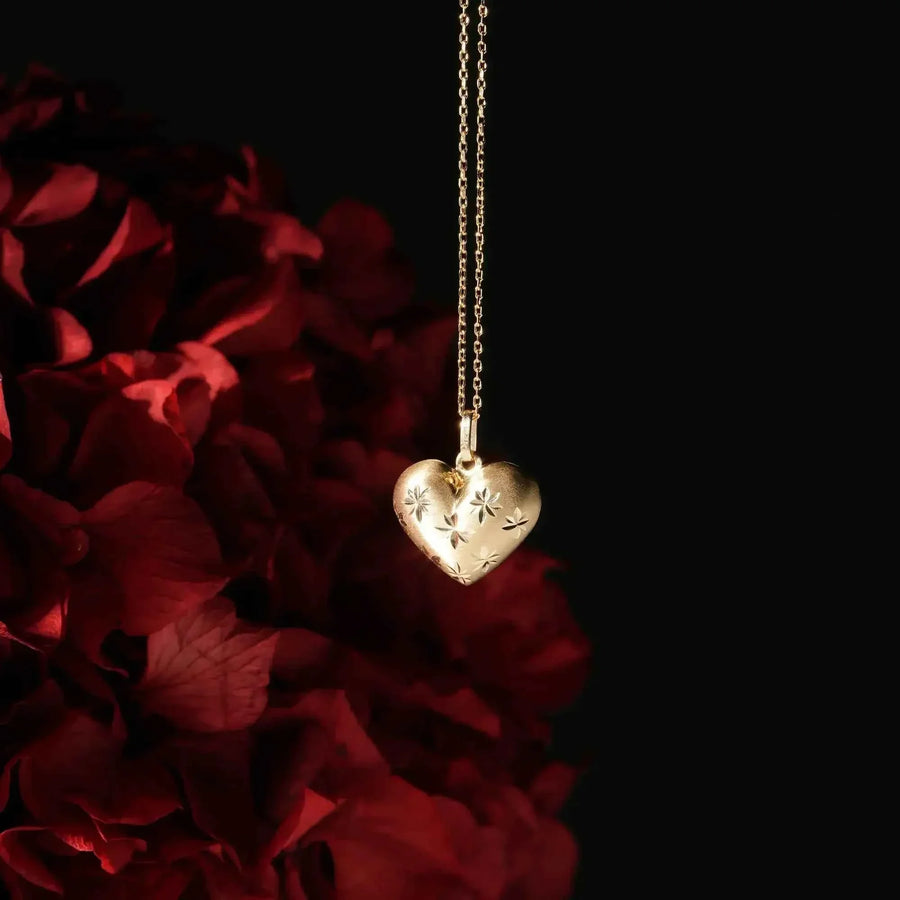 Collana cuore bombato con stella incisa in oro giallo 14kt appesa sopra petali di rosa rossa su sfondo nero
