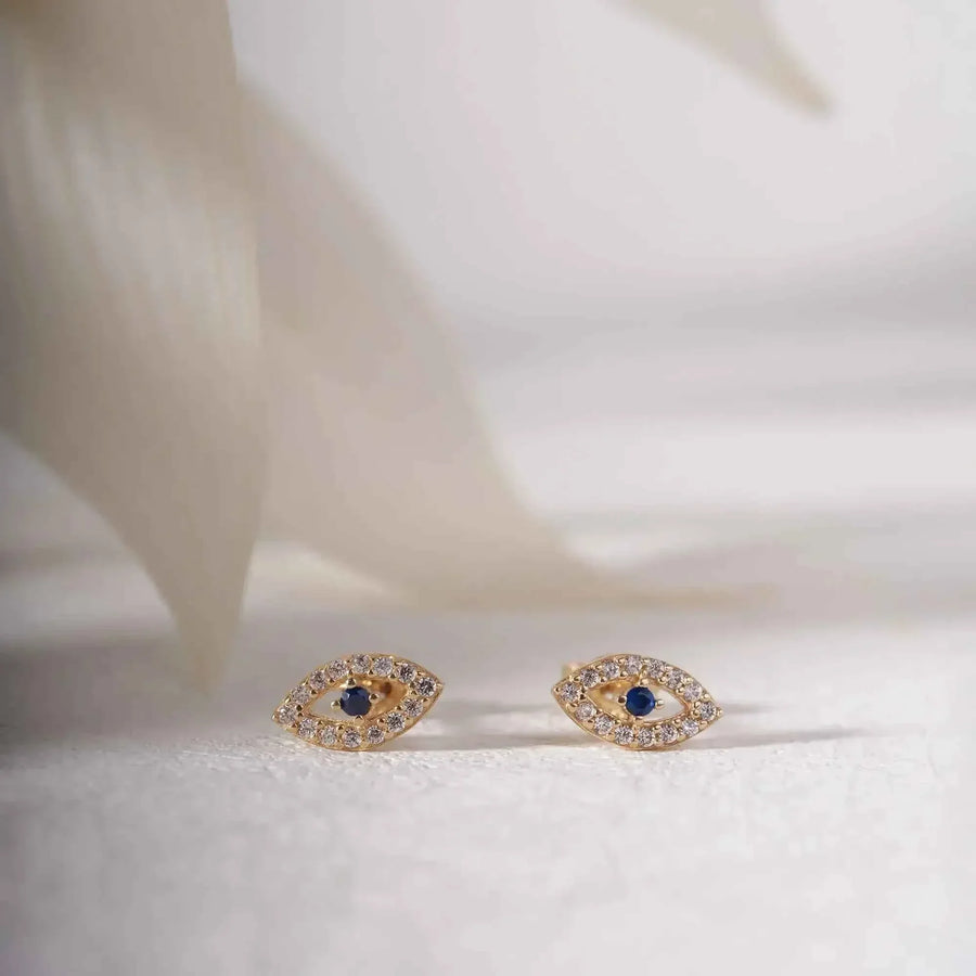 Coppia di orecchini a lobo occhio turco in oro 14kt su tessuto bianco accanto a petali delicati
