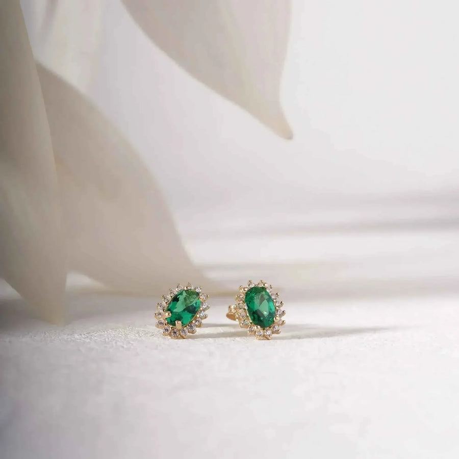 Paire de puces d'oreilles halo émeraude de synthèse en or jaune 14 carats sur un tissu blanc près de pétales délicats