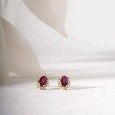 Paire de puces d'oreilles halo rubis de synthèse en or 14 carats sur un tissu blanc près de pétales délicats
