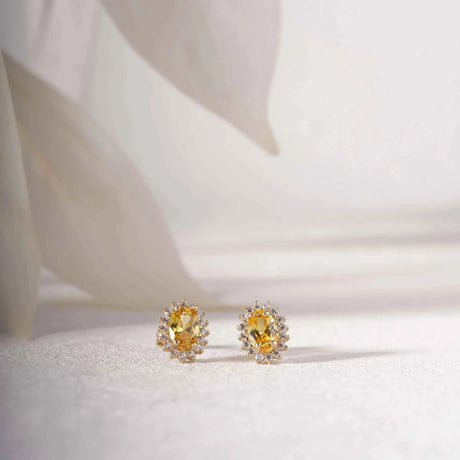 Paire de puces d'oreilles halo citrine de synthèse en or jaune 14 carats sur un tissu blanc près de pétales délicats