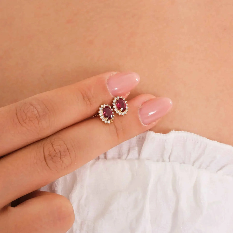 Puces d'oreilles halo rubis de synthèse en or 14 carats tenues du bout des doigts contre un haut blanc