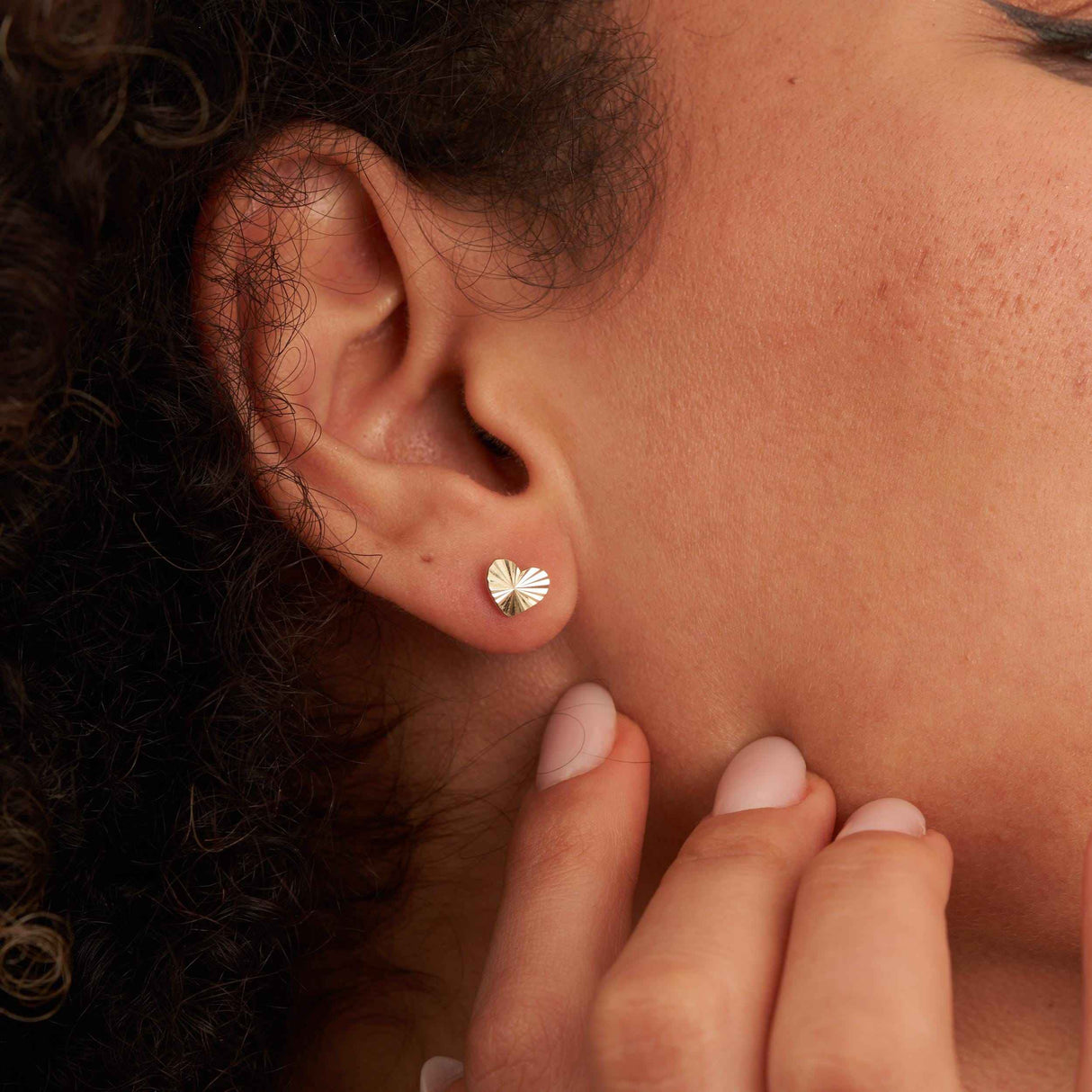 Earlobe close-up of a 14K yellow gold diamond-cut heart stud earring, fingers at the jaw