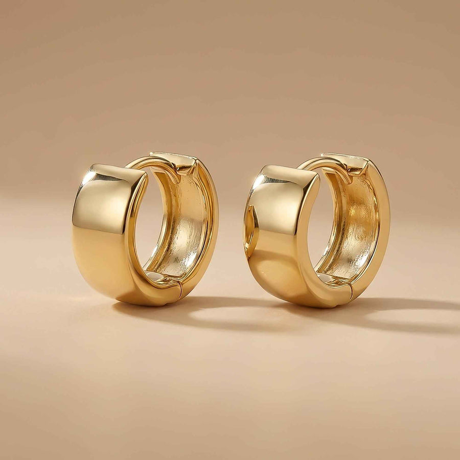 Orecchini huggie spessi in oro giallo 14kt in piedi su sfondo beige