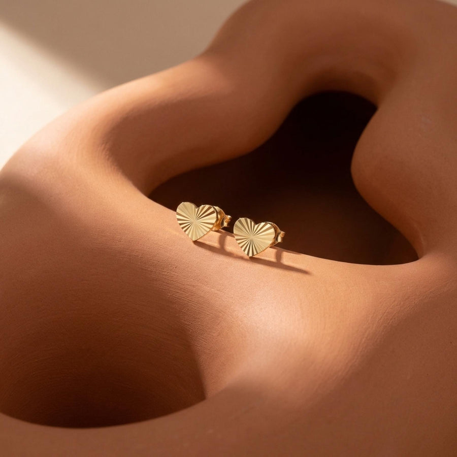 14K yellow gold diamond-cut heart stud earrings resting on a sculpted sand-colored form