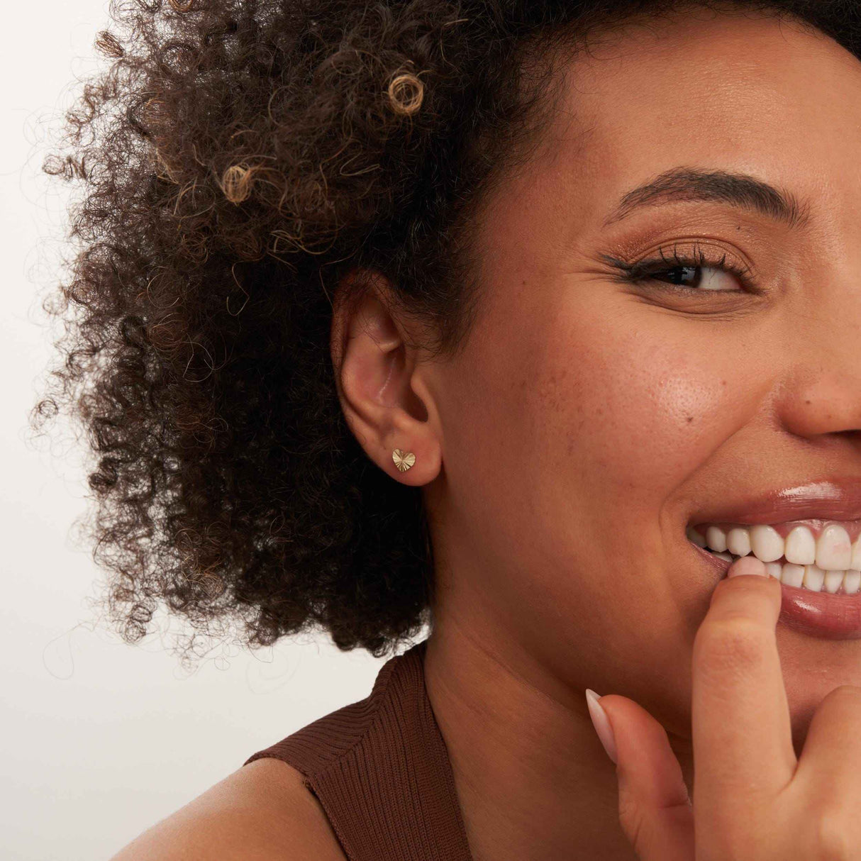 Smiling model with finger at lip wearing 14K yellow gold diamond-cut heart stud earrings