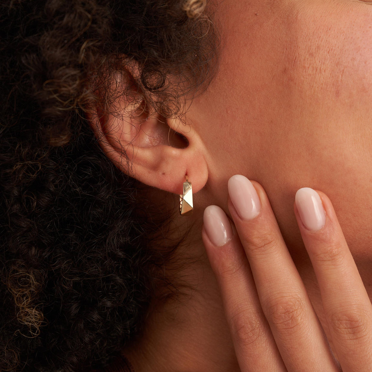 Gros plan sur une oreille avec une créole hexagonale facettée en or jaune 14 carats portée, la main sur la joue