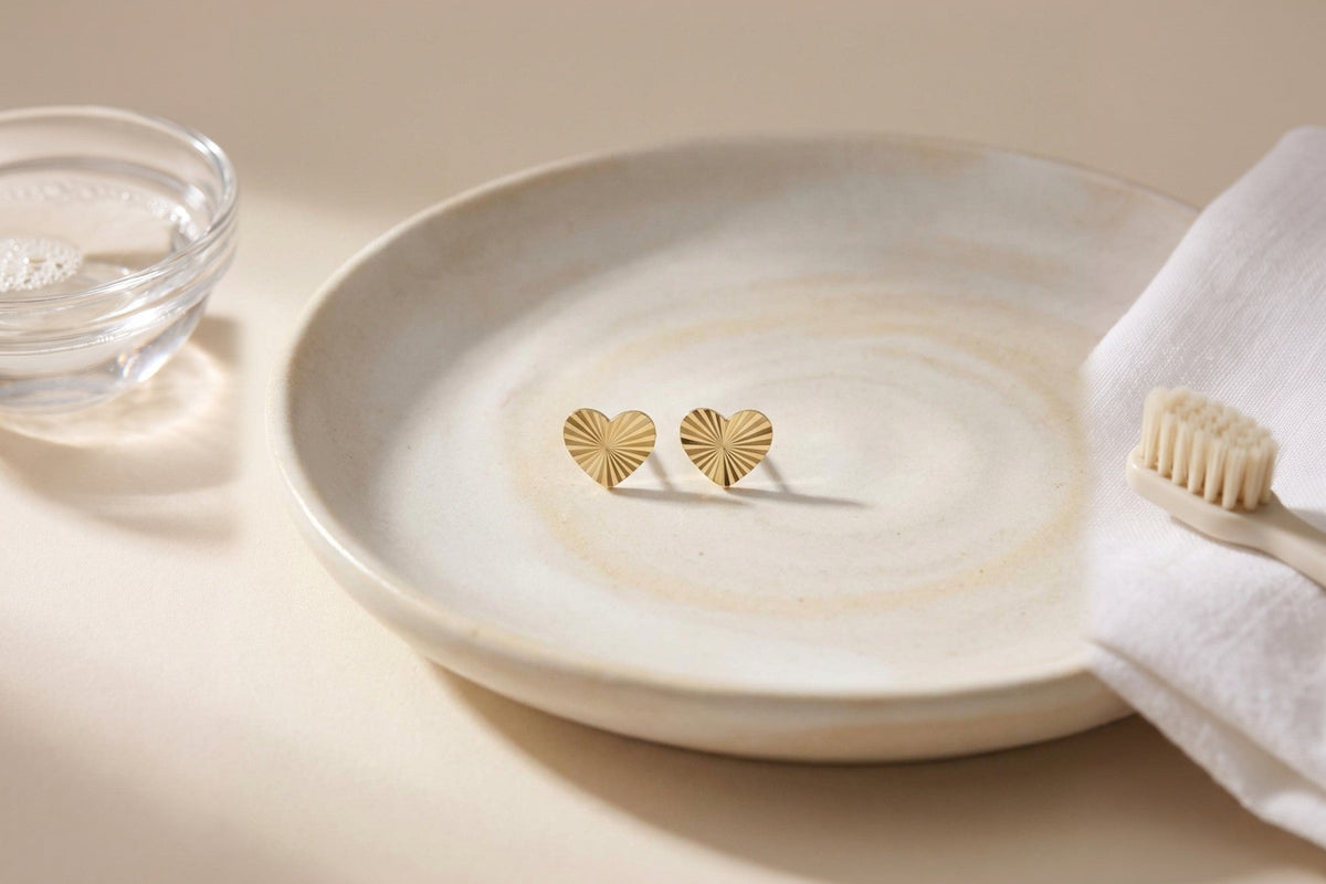 Heon 14K gold heart stud earrings in a ceramic dish beside warm soapy water, a soft cloth and a soft-bristled brush