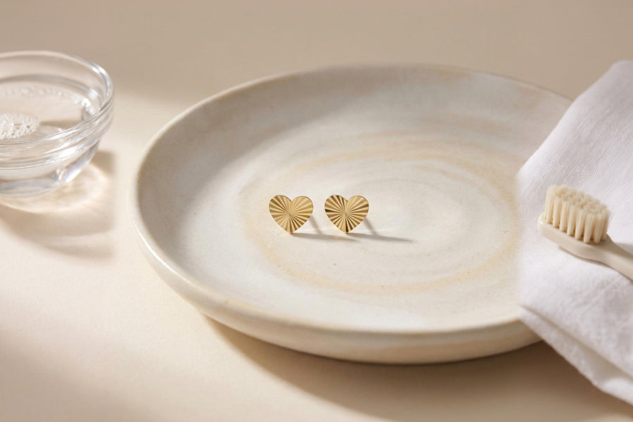 Heon 14K gold heart stud earrings in a ceramic dish beside warm soapy water, a soft cloth and a soft-bristled brush