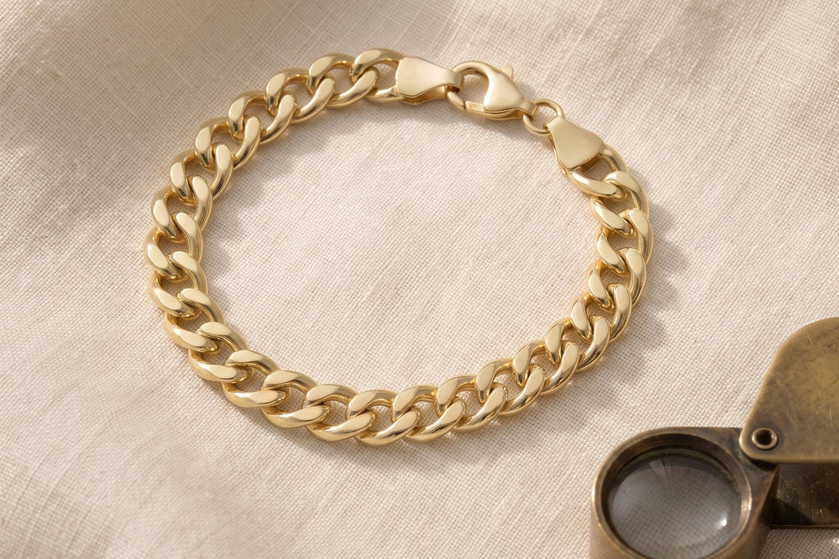 Heon 14K gold Cuban link bracelet on ivory linen beside a brass jeweler’s loupe