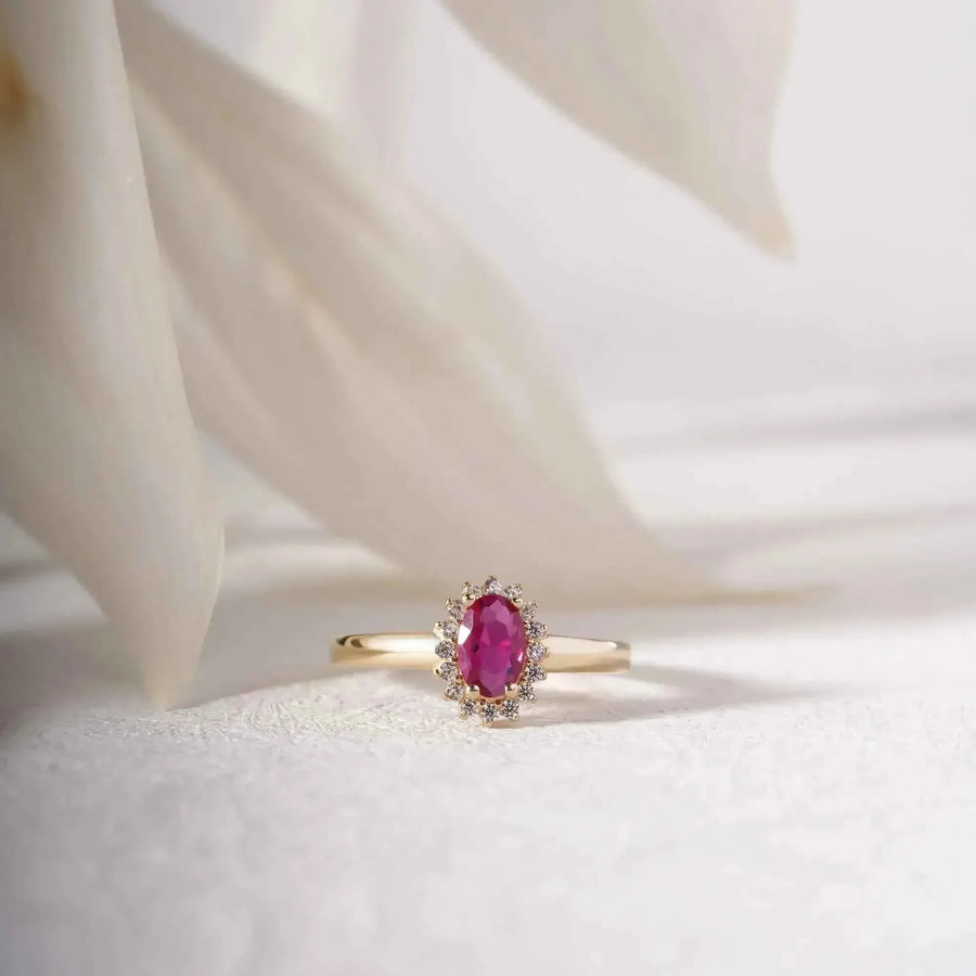 Bague pierre de naissance halo rubis de synthèse en or jaune 14 carats posée debout sur un tissu blanc près de pétales délicats