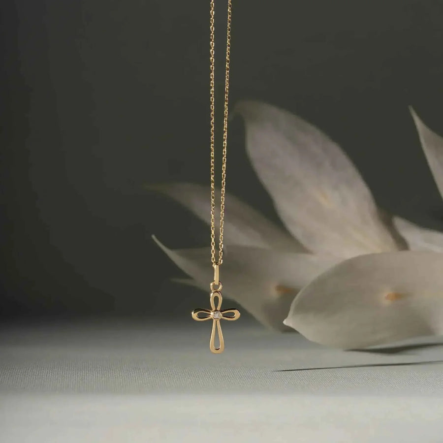 Collier pendentif croix ajourée en or jaune 14 carats suspendu devant des feuilles pâles sur fond gris