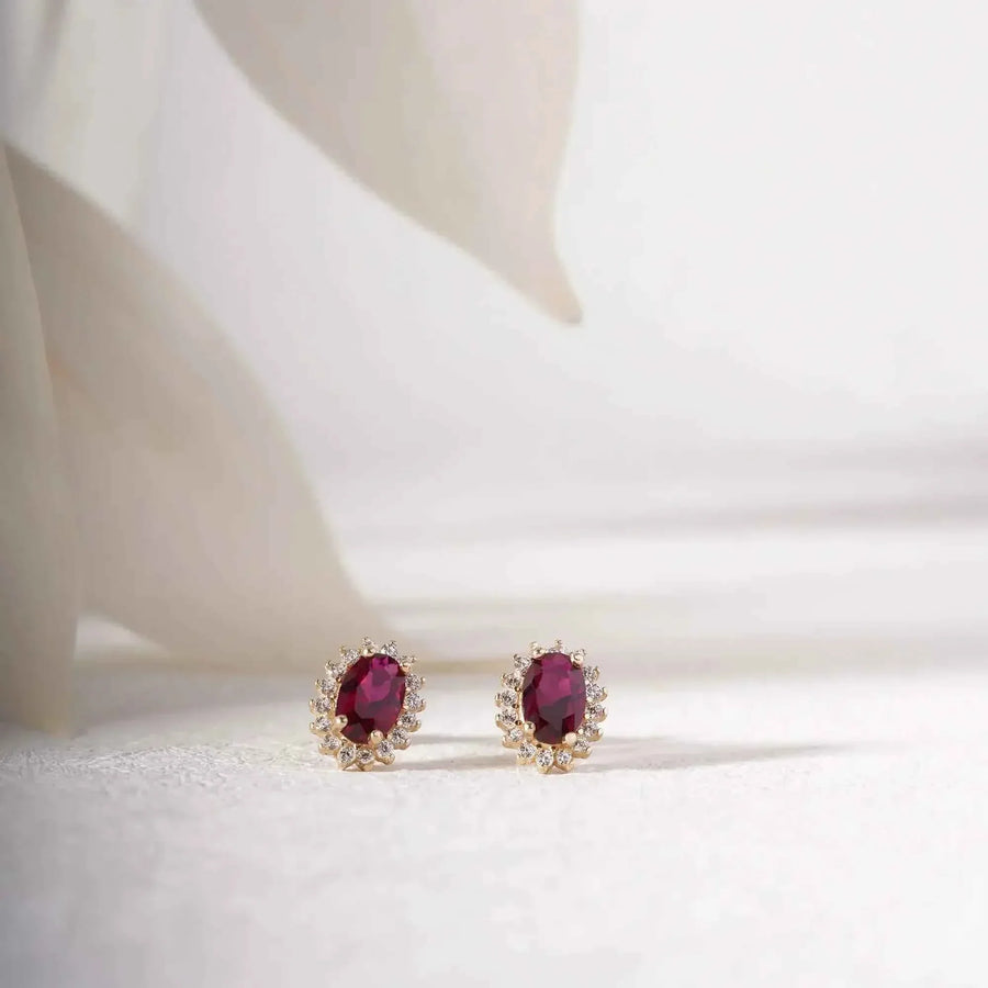 Paire de puces d'oreilles halo rubis de synthèse en or 14 carats sur un tissu blanc près de pétales délicats
