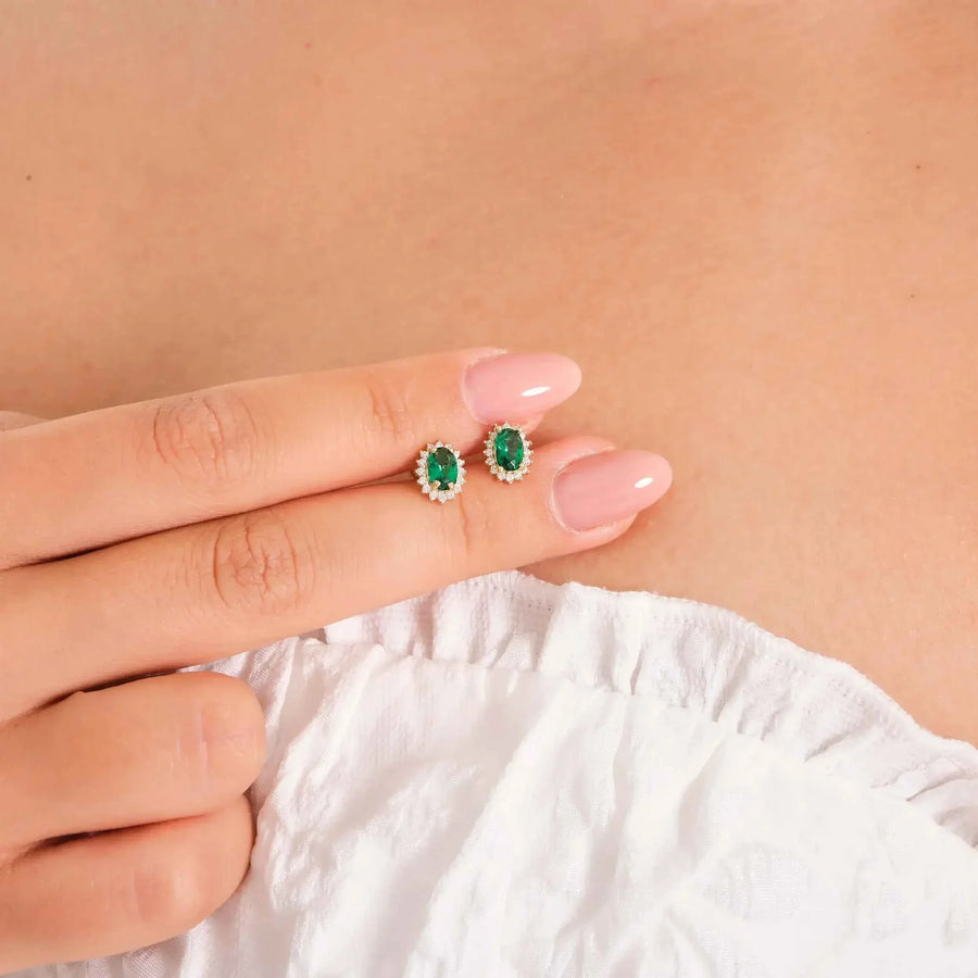 Puces d'oreilles halo émeraude de synthèse en or jaune 14 carats tenues du bout des doigts contre un haut blanc
