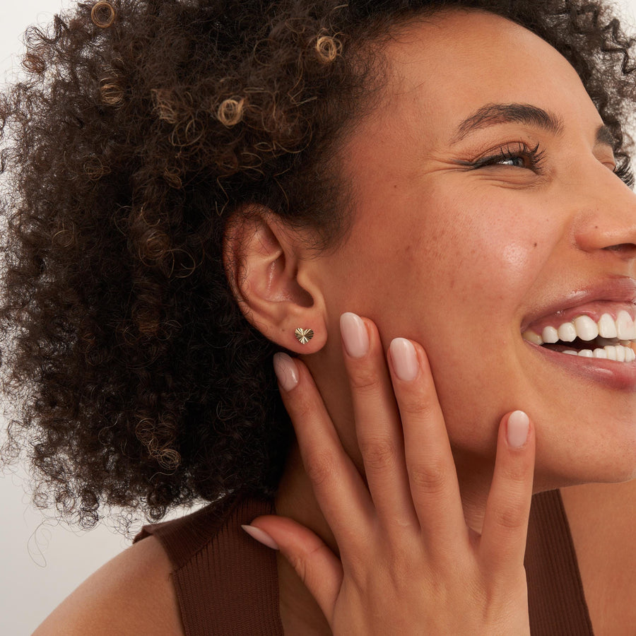 Laughing model with hand at cheek wearing 14K yellow gold diamond-cut heart stud earrings