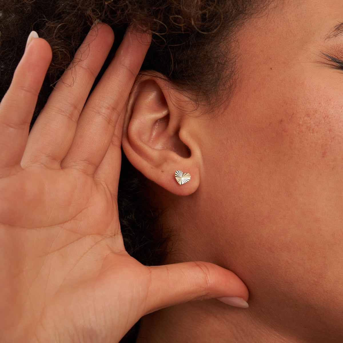 Hand cupping an ear that wears a 14K yellow gold diamond-cut heart stud earring