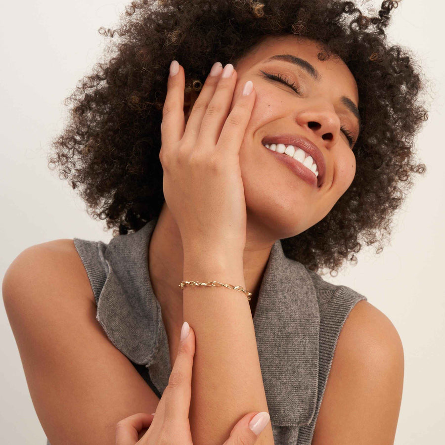 Modelo sonriente con la mano en la mejilla y una pulsera de cadena retorcida de eslabones infinito en oro amarillo de 14 quilates