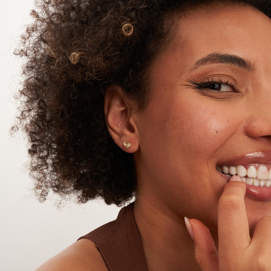 Smiling model with finger at lip wearing 14K yellow gold diamond-cut heart stud earrings
