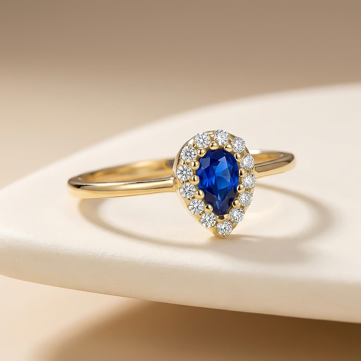 14K yellow gold pear halo ring with lab-grown sapphire on beige backdrop