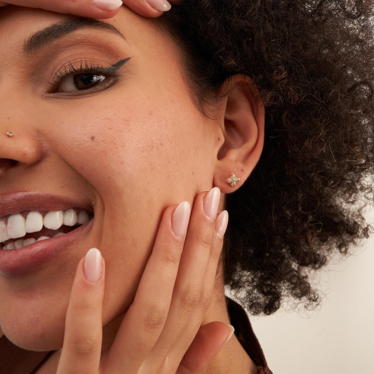 Mannequin souriant à l'objectif, main sur la joue, portant des puces d'oreilles fleur en oxyde de zirconium et or jaune 14 carats
