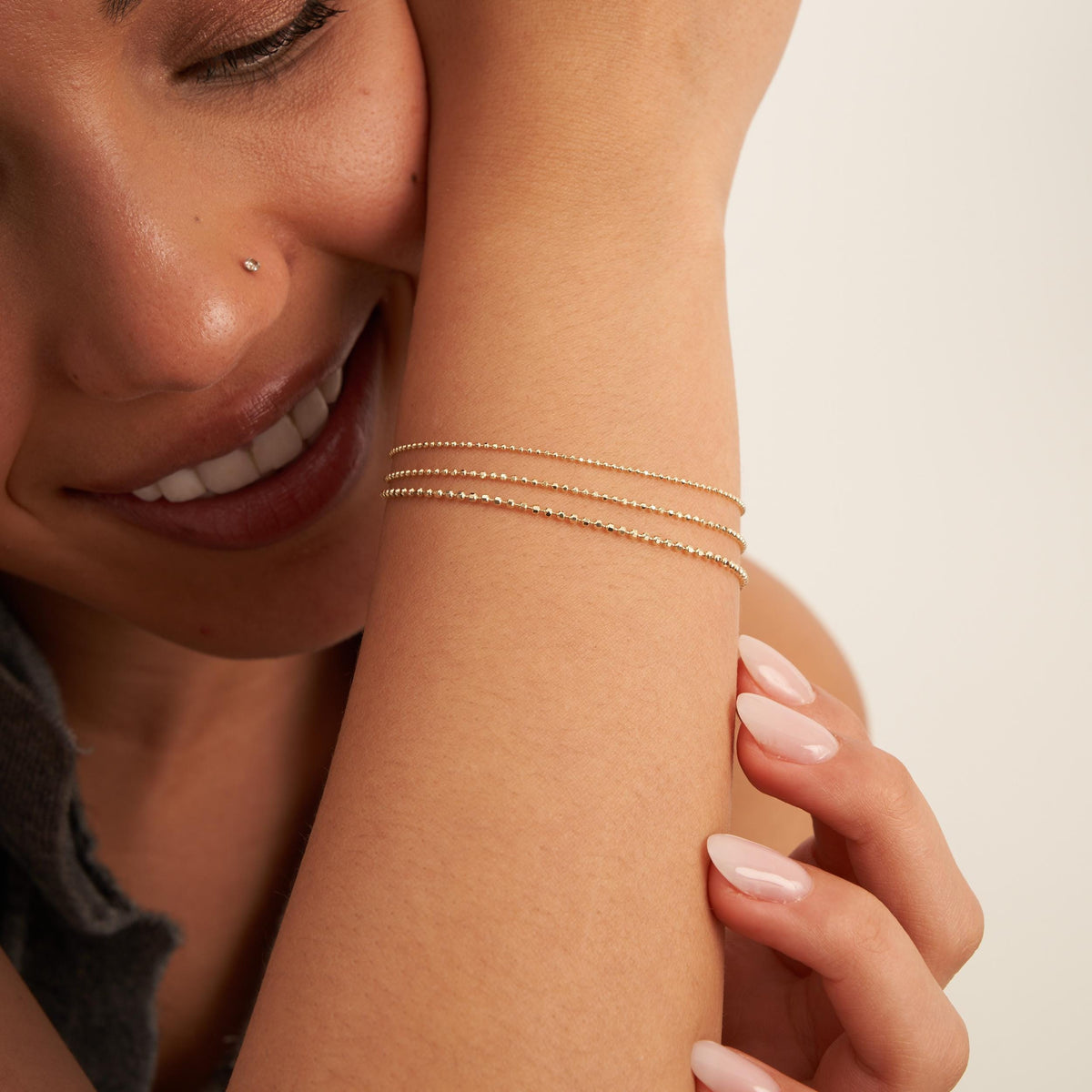 La modelo sonriente apoya la mejilla en la muñeca, con pulseras de cadena de bolitas en oro amarillo de 14 quilates apiladas